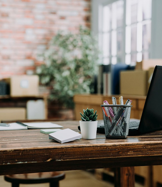 Organized workspace with a laptop, potted plant, pens in a holder on a wooden desk; brick wall in the background.