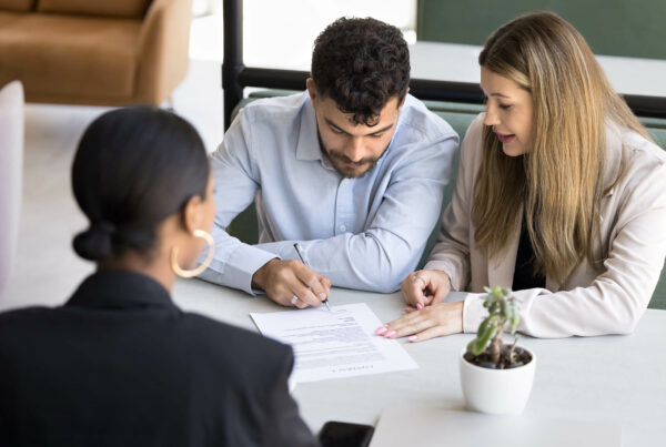 Three professionals reviewing and signing a document at a table with a small plant in the foreground.