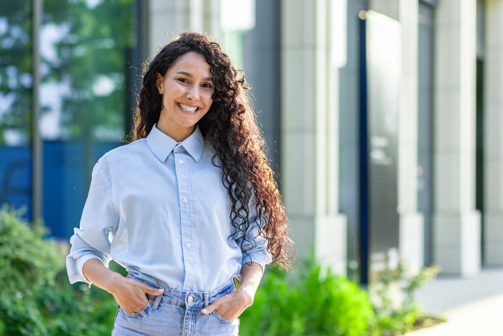 Smiling person in a light blue shirt standing outdoors, hands in pockets, with greenery and a modern building in the backgrou
