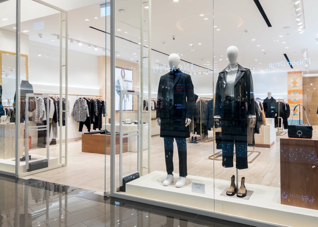 Fashion store window display with mannequins dressed in winter coats. Inside, various garments are hung on racks.