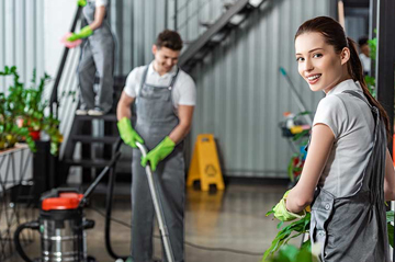 Workers cleaning an indoor space, with one smiling at the camera. They wear gloves and use vacuums and mops.