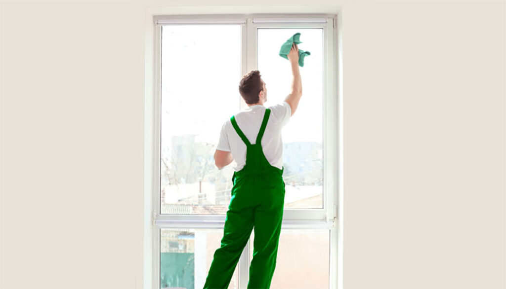 Person in green overalls cleaning a large window with a cloth, standing indoors with city view outside.