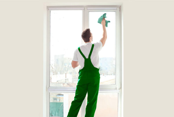Person in green overalls cleaning a large window with a cloth, standing indoors with city view outside.