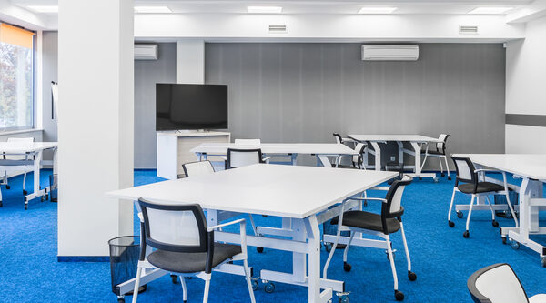Broad view of interior learning classroom that has blue carpets, white desks and chairs and a whiteboard for teaching.