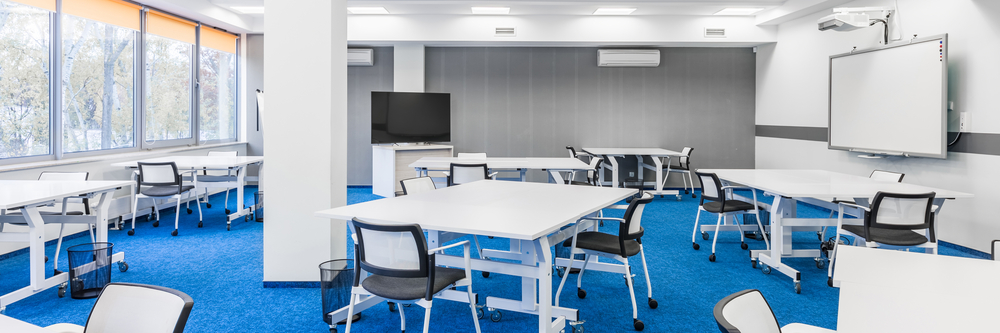 Broad view of interior learning classroom that has blue carpets, white desks and chairs and a whiteboard for teaching.
