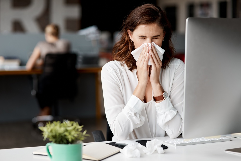 Young woman in an office who is sick and blowing her nose sitting at her desk.